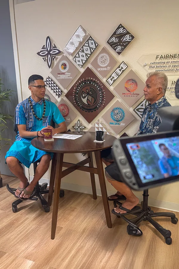 Two men in traditional Pacific Islander attire are being interviewed in front of a wall with cultural symbols. One man is wearing an "ASCC Cafe" mug. The interview is part of the "About Us" content.