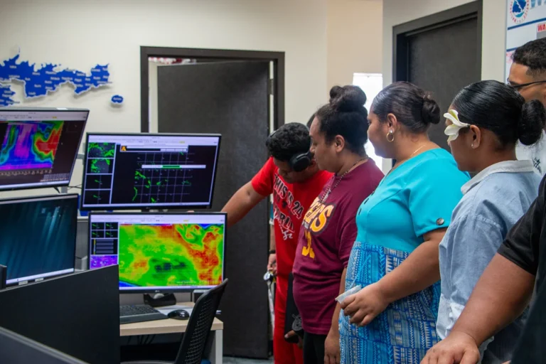 A group of people stand in front of computer screens displaying weather data and maps, likely discussing weather patterns. A wall map of islands is behind them. This image is featured on the ANA Pacific Region website.