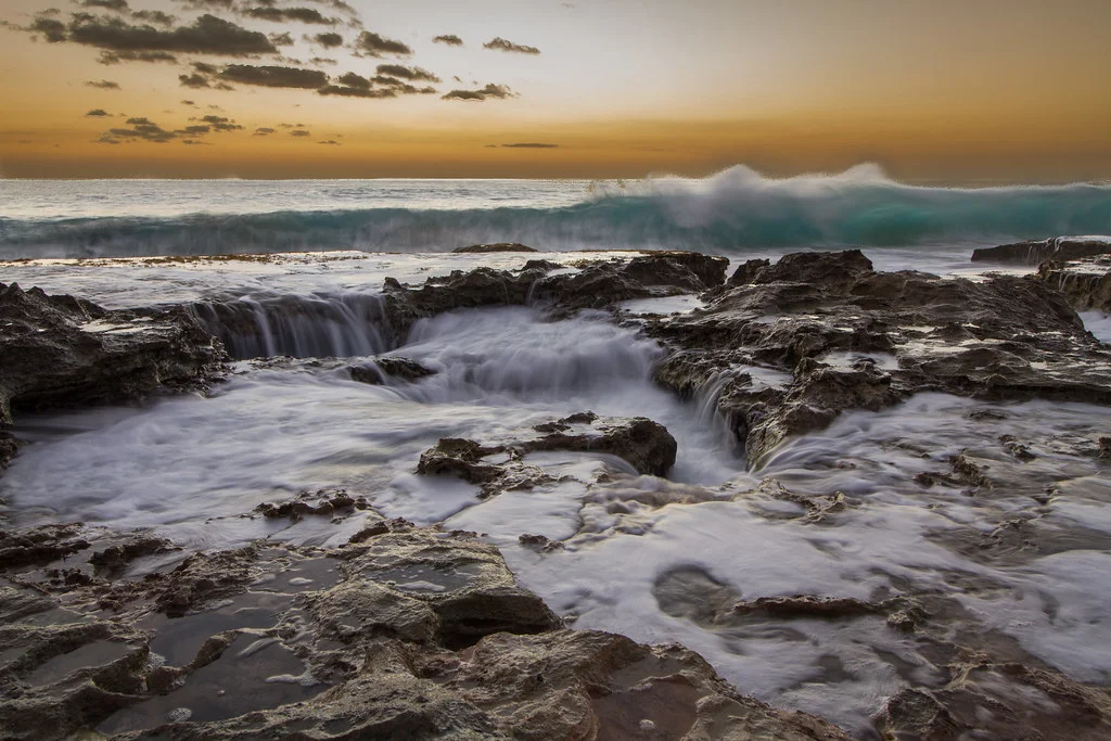 Waves crash over rocky shore at sunset, creating waterfalls and sea foam. Golden light illuminates the sky and turbulent water, capturing a dramatic coastal scene. Photo by Jeff Chun-Lum.