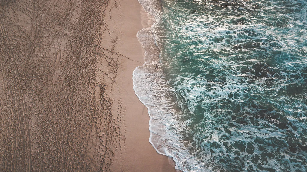 Aerial view of a sandy beach where the waves meet the shore. Footprints are visible in the sand, suggesting a peaceful beach vacation along the coastline. Photo by Jeff Chun-Lum.