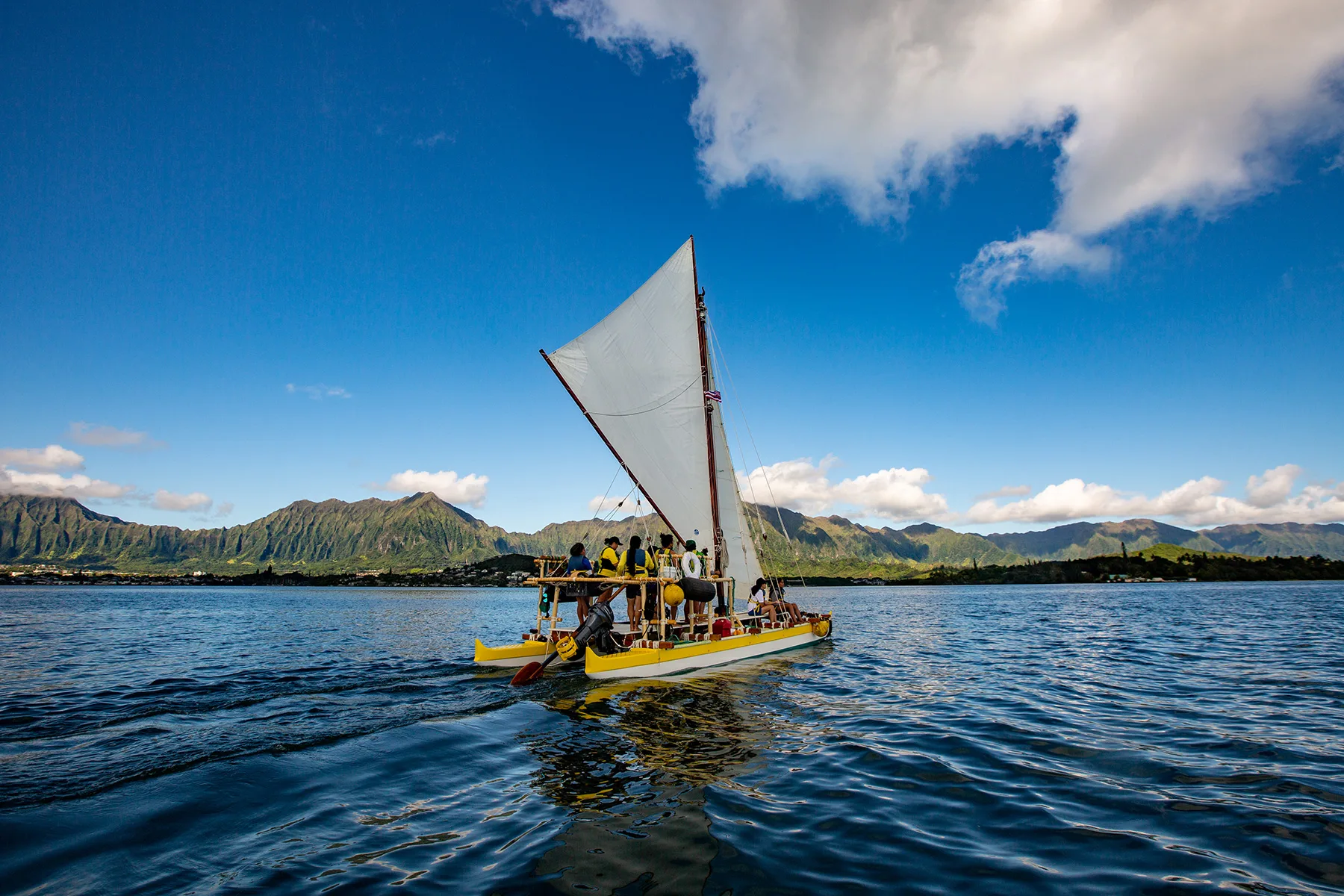 Outrigger sailing canoe glides across Kaneohe Bay, Hawaii, with lush green mountains in the background under a bright blue sky. People enjoy a scenic 'Home' adventure on the water. This image is featured on the ANA Pacific Region website.