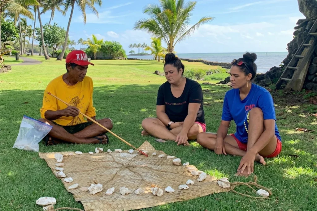 A Native Hawaiian man in a yellow shirt points to a woven mat with stones arranged on it, explaining a traditional game to two women on a grassy lawn near the ocean with palm trees. This image is featured on the ANA Pacific Region website.