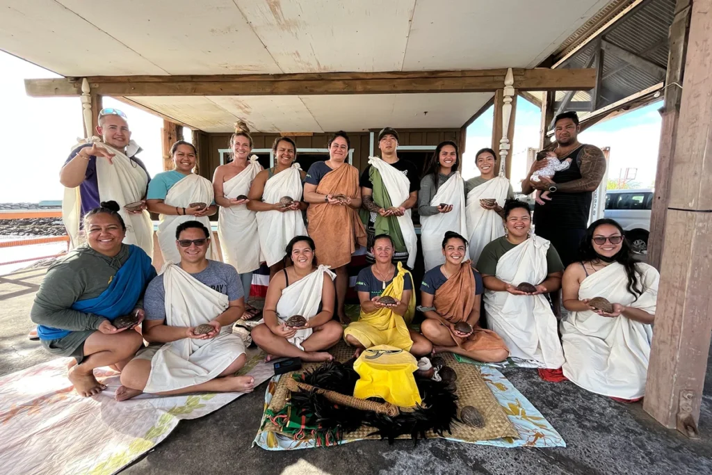 A group of people in traditional attire gather outdoors, holding coconuts. Some sit cross-legged around ceremonial items. The setting appears to be a cultural or spiritual event. This image is featured on the ANA Pacific Region website.