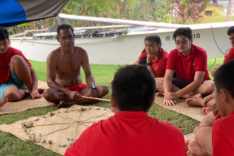 A shirtless Micronesian man sits cross-legged, teaching navigation skills to students in red shirts using a traditional star compass made of stones on a woven mat. A small sailboat is visible in the background. This image is featured on the ANA Pacific Region website.