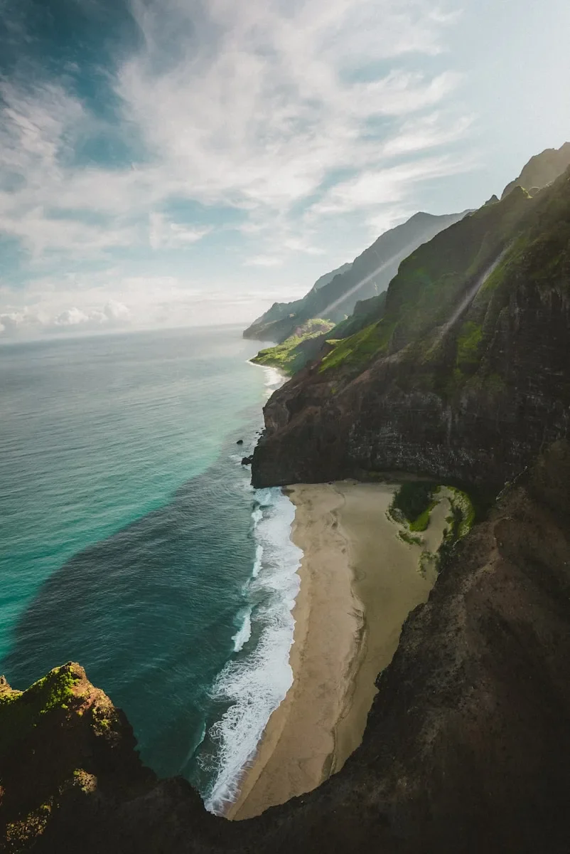 Aerial view of the Na Pali Coast in Kauai, Hawaii, showcasing dramatic green cliffs meeting turquoise ocean waters and a secluded sandy beach. Photo by Karsten Winegeart.