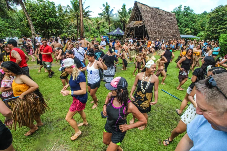 A diverse group of people in colorful attire participate in a lively outdoor festival, dancing and enjoying themselves in a tropical setting with palm trees and a thatched-roof hut. This image is featured on the ANA Pacific Region website.