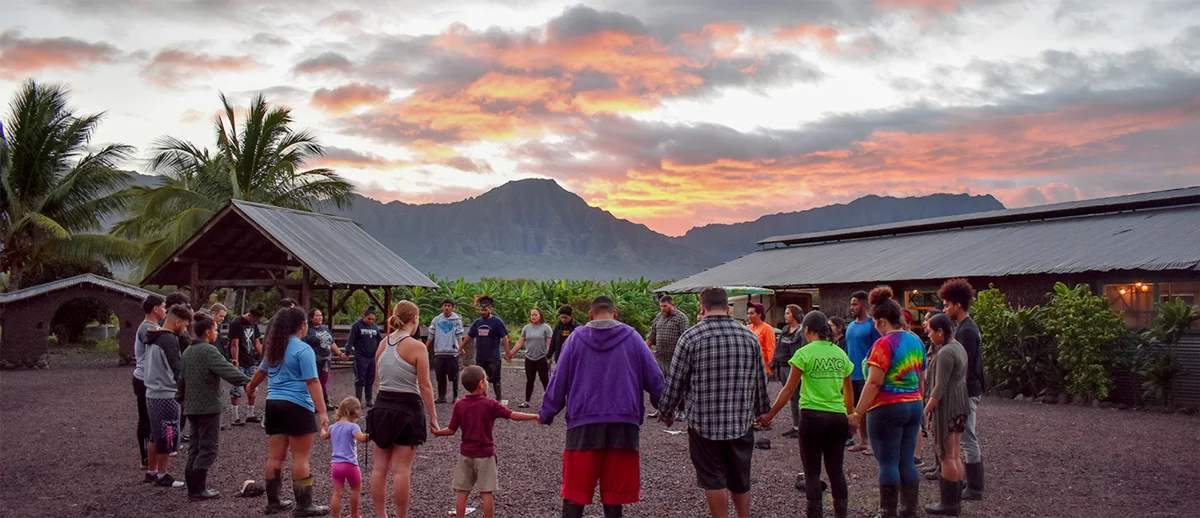 A diverse group holds hands in a circle at sunset, with mountains and palm trees in the background. The sky is a mix of orange, pink, and gray, creating a warm atmosphere. This image is featured on the ANA Pacific Region website.