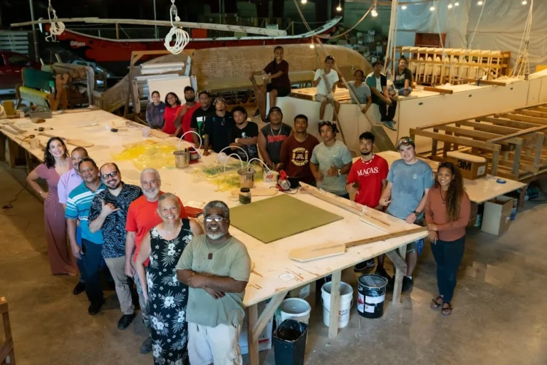 A diverse group of people, including men, women, and children, stand together around a large wooden table in a workshop, smiling and posing for a photo. Various boat-building materials and tools are visible in the background, suggesting a community boat-building project. This image is featured on the ANA Pacific Region website.